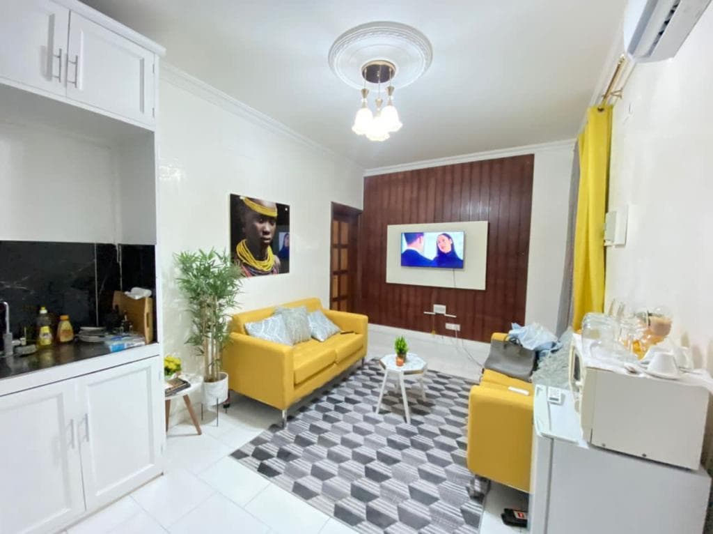 A living area features a yellow sofa and matching armchairs, complemented by a geometric area rug. A small coffee table is situated in the center, while a flat-screen TV is mounted on the wall. A kitchenette is visible, equipped with basic appliances.