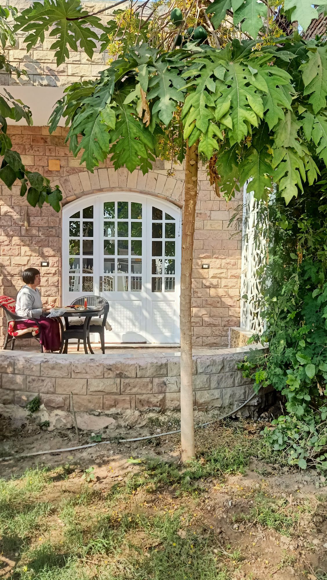 A patio area is visible, featuring a round black table and two chairs, partially shaded by a small tree. The stone wall of the building is characterized by a large, arched window that allows light to enter. Lush greenery surrounds the space.