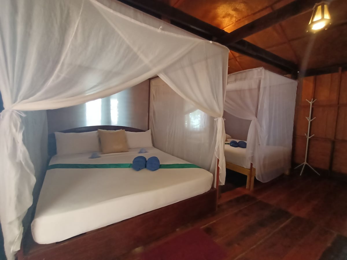 Two spacious beds are nestled in a rustic wooden interior, each surrounded by sheer white canopies. Soft towels are neatly arranged at the foot of each bed, offering a sense of comfort. Natural light filters through small windows, enhancing the welcoming ambiance.