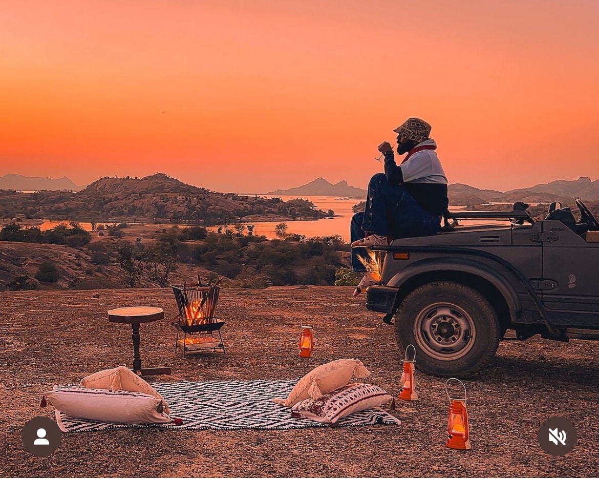 A serene outdoor setting is depicted with a figure seated on a vehicle, enjoying the sunset over distant hills and water. A picnic area is set with a patterned mat, cushions, and glowing lanterns, creating a tranquil atmosphere for relaxation.