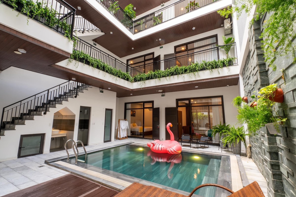 A central pool area is framed by three levels of balconies adorned with greenery. A pink inflatable flamingo floats in the inviting water. Stairs lead to upper floors, while large windows provide views into the surrounding spaces.