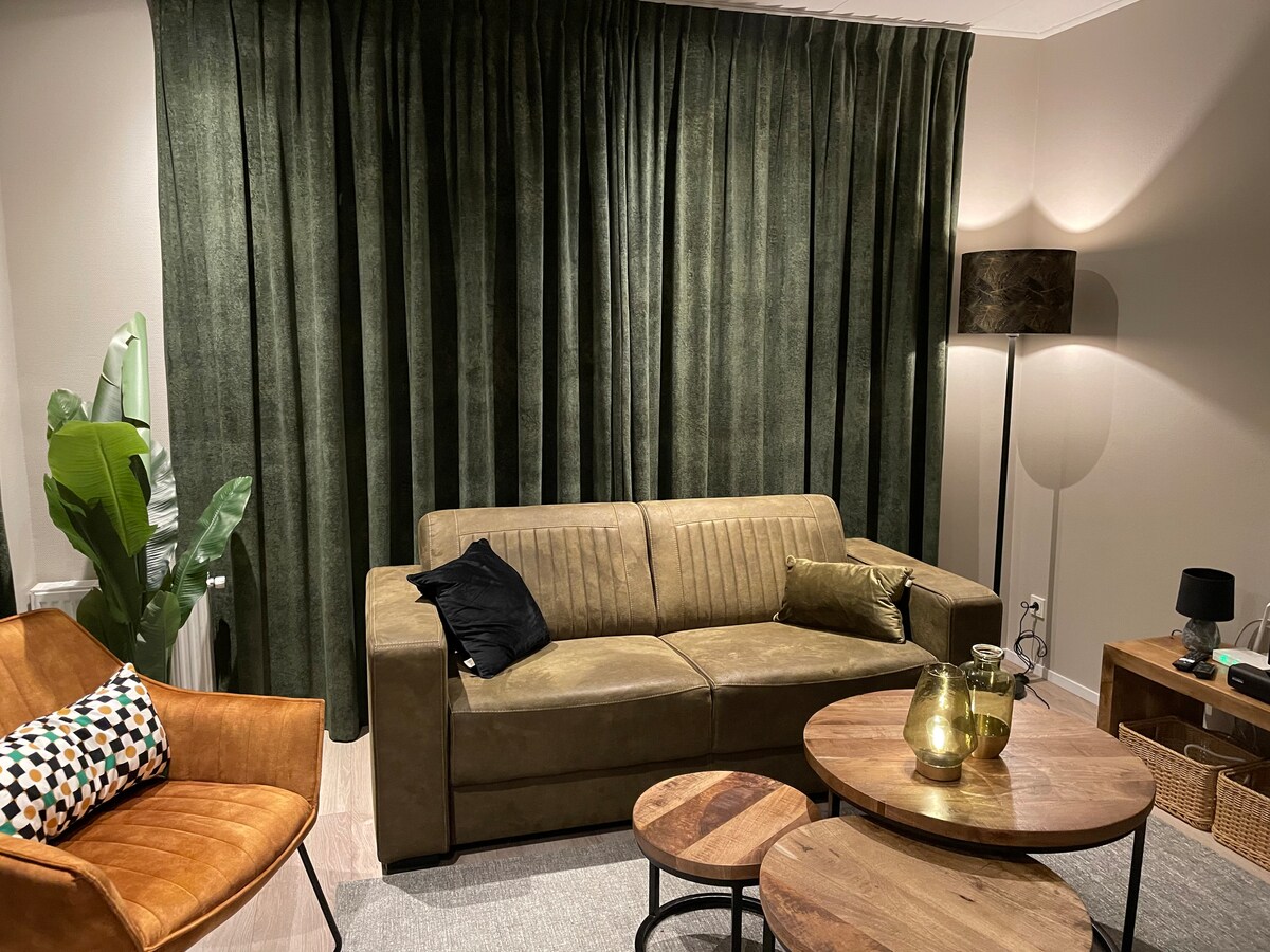 A cozy living area features a soft green color palette, with a comfortable sofa and an armchair. Natural light is softly diffused by the dark green curtains. Two round wooden tables are positioned in front of the seating, complemented by a decorative lamp and houseplants.