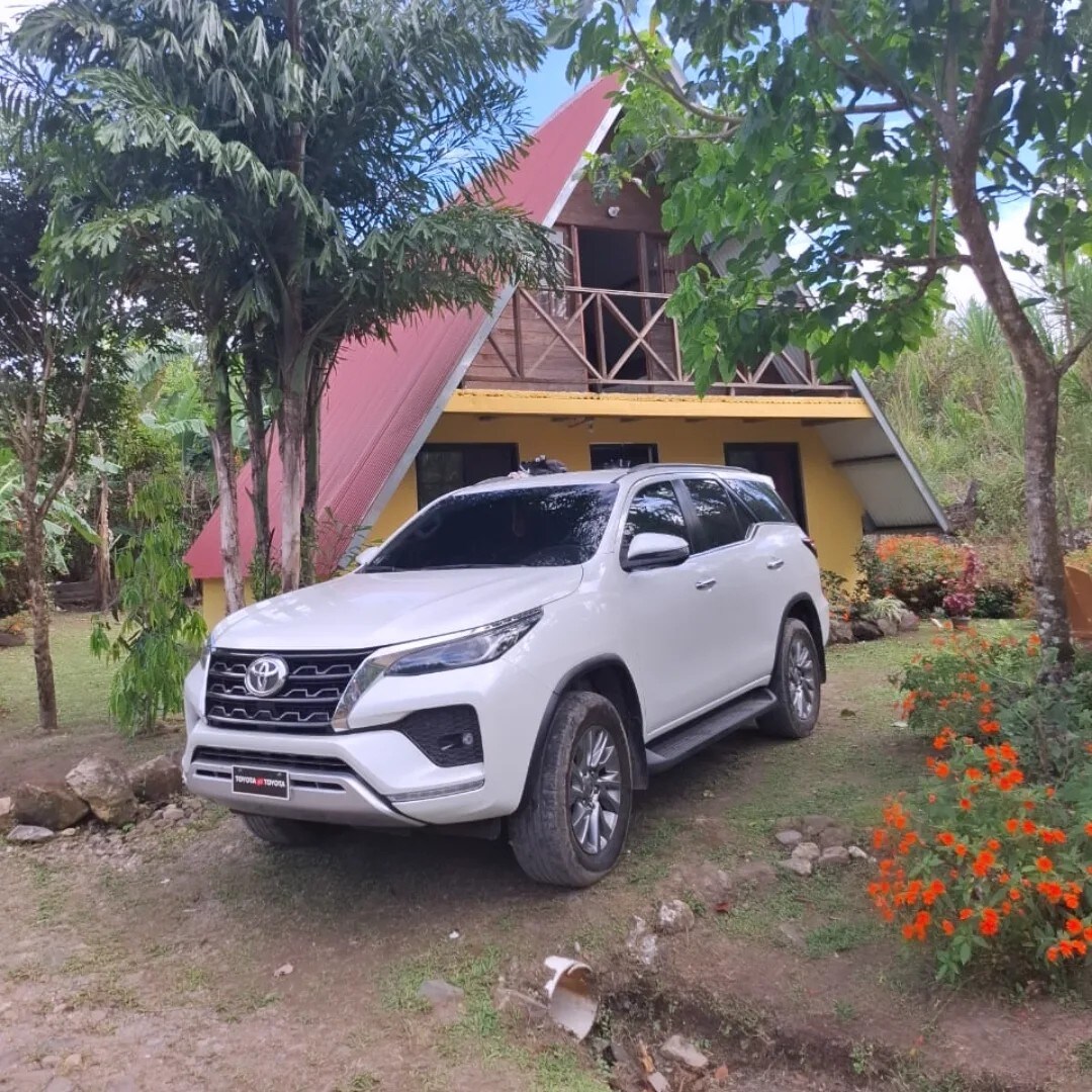 A white SUV is parked in front of a triangular-roofed cabin, surrounded by lush greenery and vibrant flowers. The cabin features a mix of yellow and brown tones, with a spacious porch and wooden accents visible.