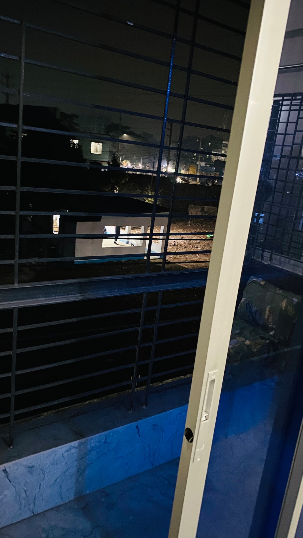 A nighttime view from a balcony is presented, framed by a securely fastened door. The balcony overlooks a well-lit neighborhood, with lights from distant buildings twinkling against the dark backdrop of the evening sky.
