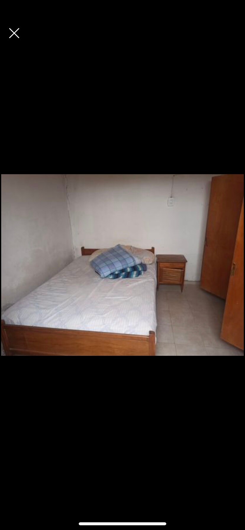 A simple sleeping area features a wooden bed with a light-colored mattress and a plaid throw pillow. A small nightstand is situated beside the bed, while a wooden closet is visible against the wall. Soft natural light enters through a nearby window.