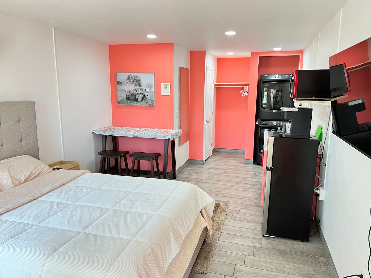 A spacious suite features light-colored walls accented with coral hues. A comfortable queen bed is positioned to one side. A large desk with stools, a refrigerator, and a compact kitchen area are visible. Natural light is provided by overhead lighting, while images and furnishings add character.