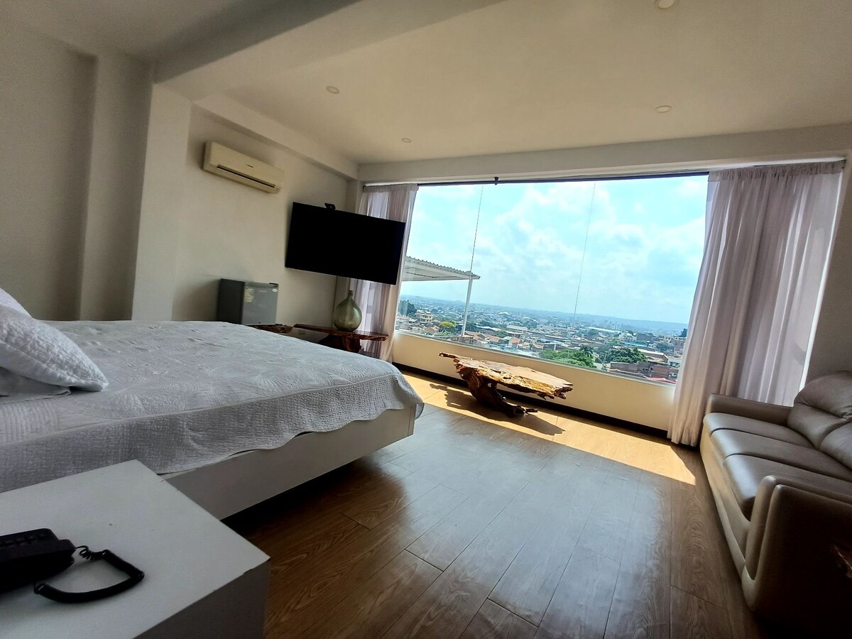 A spacious room is highlighted by a large window offering panoramic views of the city. The bed is positioned near the window, while a comfortable sofa and a modern entertainment unit are present in the space. Natural light floods the room through sheer curtains.