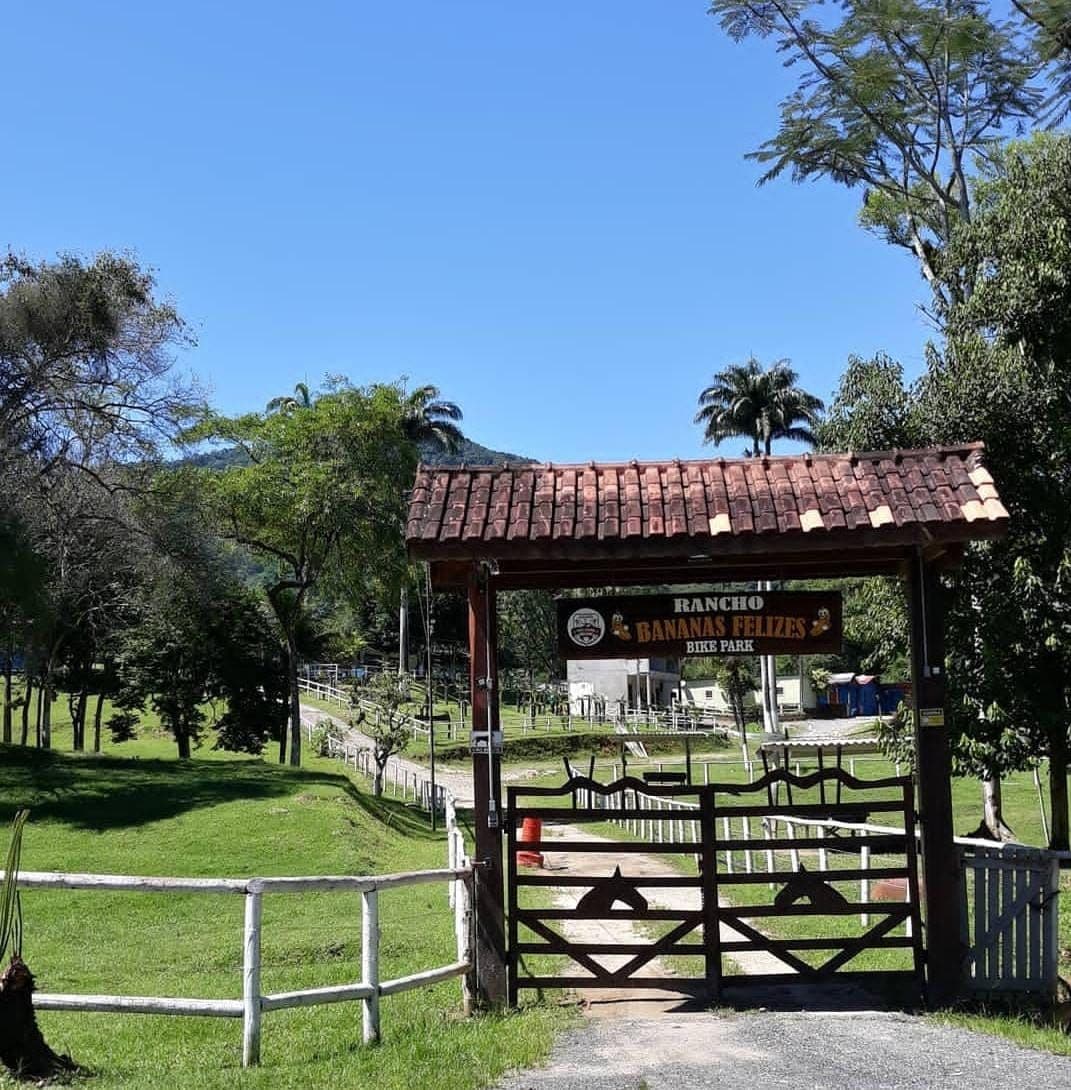 The entrance of Rancho Bananas Felizes is framed by a rustic wooden gate, embellished with a welcoming sign. Lush greenery surrounds the area, extending towards the horizon, where hills and trees are visible under a clear blue sky.