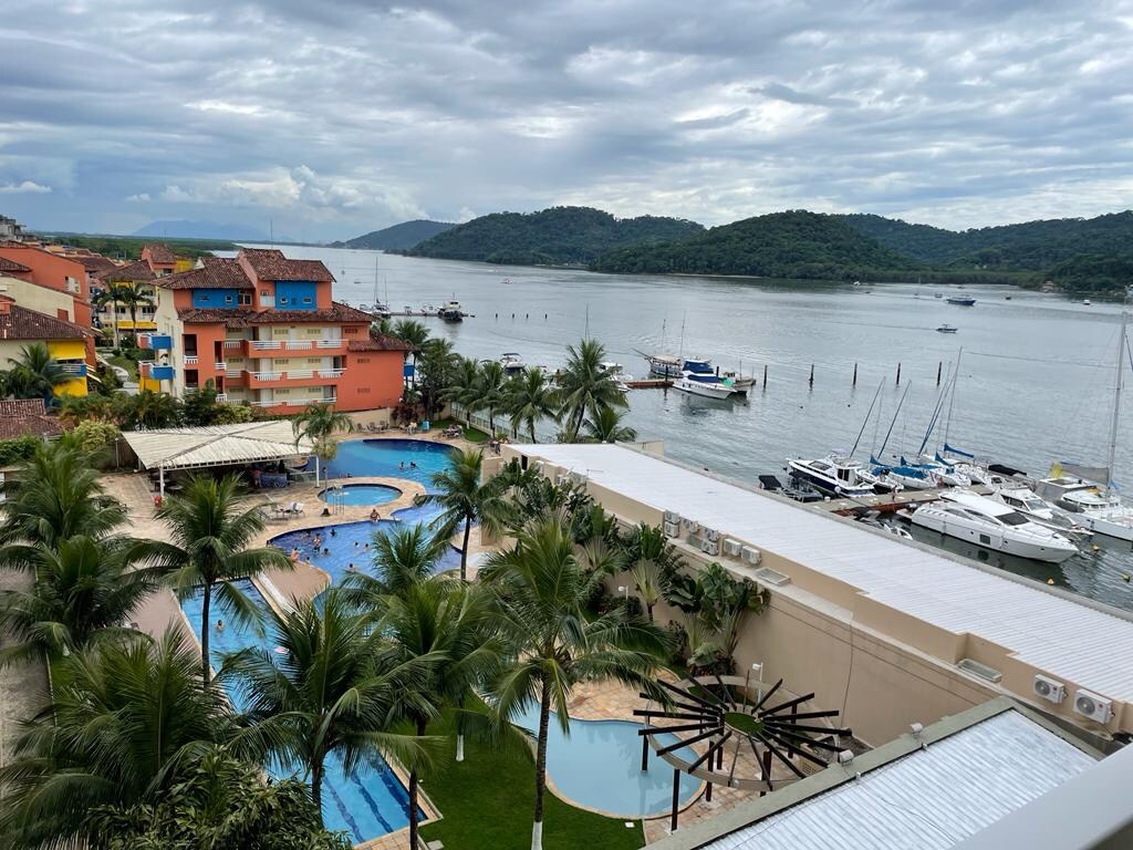 A scenic view of a waterfront resort is displayed, featuring multiple swimming pools surrounded by tropical landscaping. Boats are visible along the water, with mountains in the background under a cloudy sky.