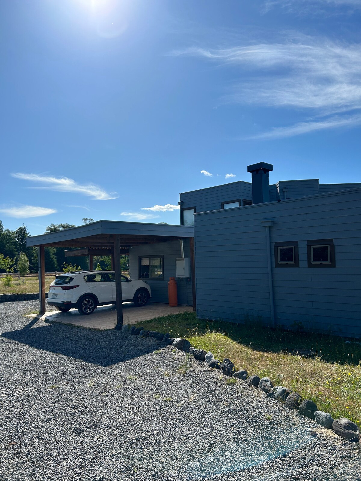 A modern two-story building is depicted under a clear blue sky. A carport shades a vehicle in front of the blue exterior. Gravel pathways and green spaces surround the property, with well-defined landscaping elements enhancing the overall setting.