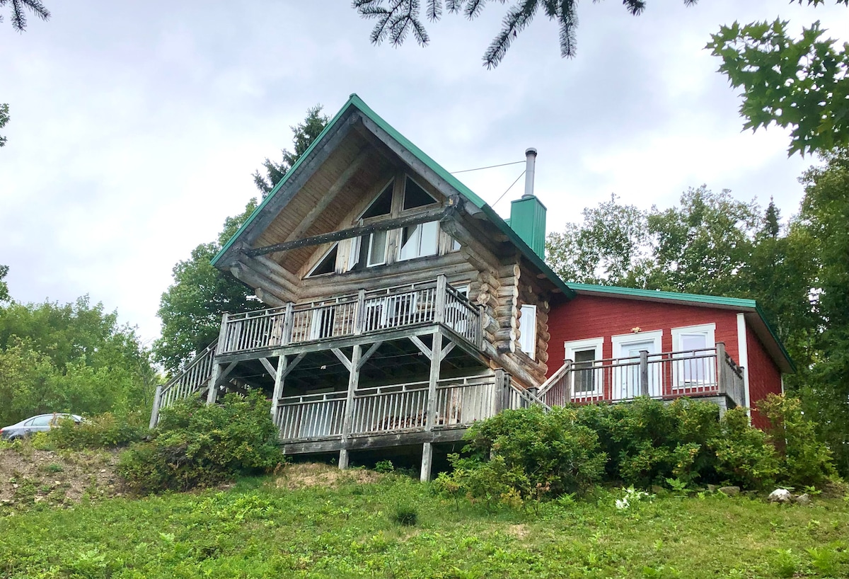 A charming log cabin is positioned on a hillside, featuring a sloped roof and multiple balconies. A red section of the building contrasts with the natural surroundings, while lush greenery frames the cabin, enhancing its natural setting.