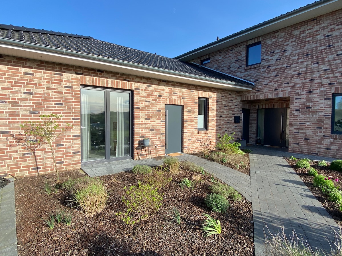 The exterior of a modern brick building is featured, showcasing a landscaped pathway with various plants and shrubs. Large glass doors offer views into the interior, while a small chair is positioned nearby. A clear blue sky complements the scene.