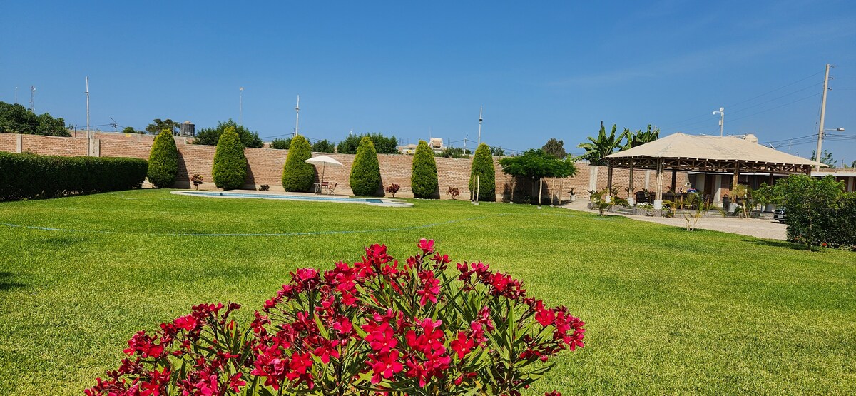 A spacious outdoor area is presented with well-manicured green grass and neatly trimmed shrubbery. Colorful flowering plants are visible in the foreground, while a shaded gazebo can be seen in the distance, surrounded by palm trees.