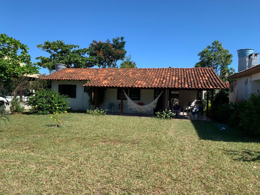 A single-story house with a terracotta roof is surrounded by a green lawn. A hammock is suspended from the porch, which provides shaded seating. Lush trees and shrubs frame the exterior, contributing to a peaceful outdoor setting.