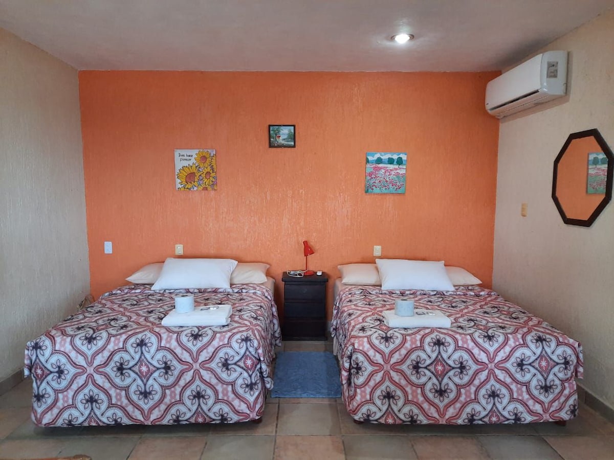 The room features two spacious beds, each adorned with colorful patterned bedding. An orange accent wall adds warmth, complemented by decorative wall art. A nightstand is positioned between the beds, and an air conditioning unit is mounted on the wall, providing comfort.
