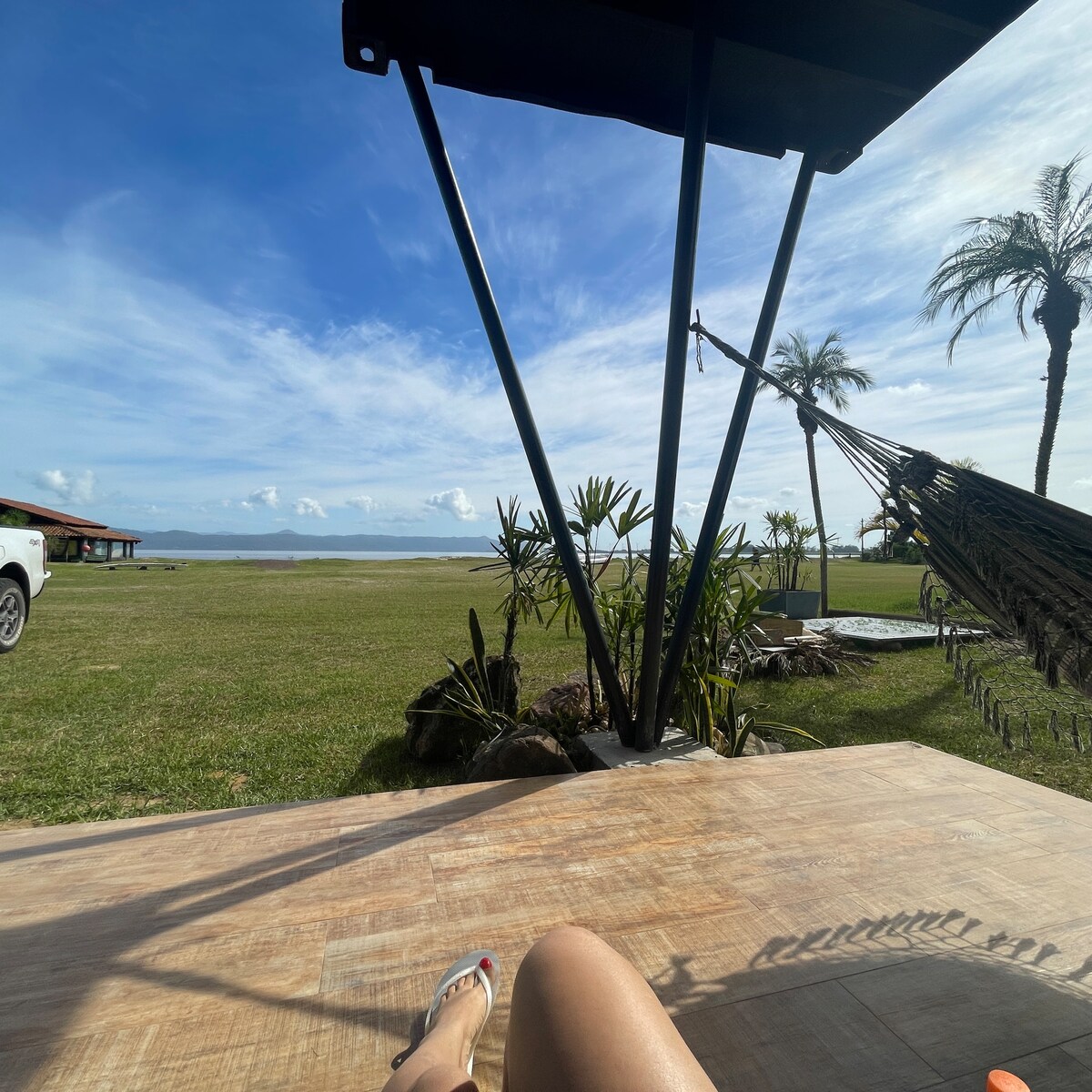A view from a shaded area showcases a large grassy expanse leading to a calm lagoon, with palm trees framed against a clear blue sky. A hammock is visible to the side, providing a relaxed outdoor space.