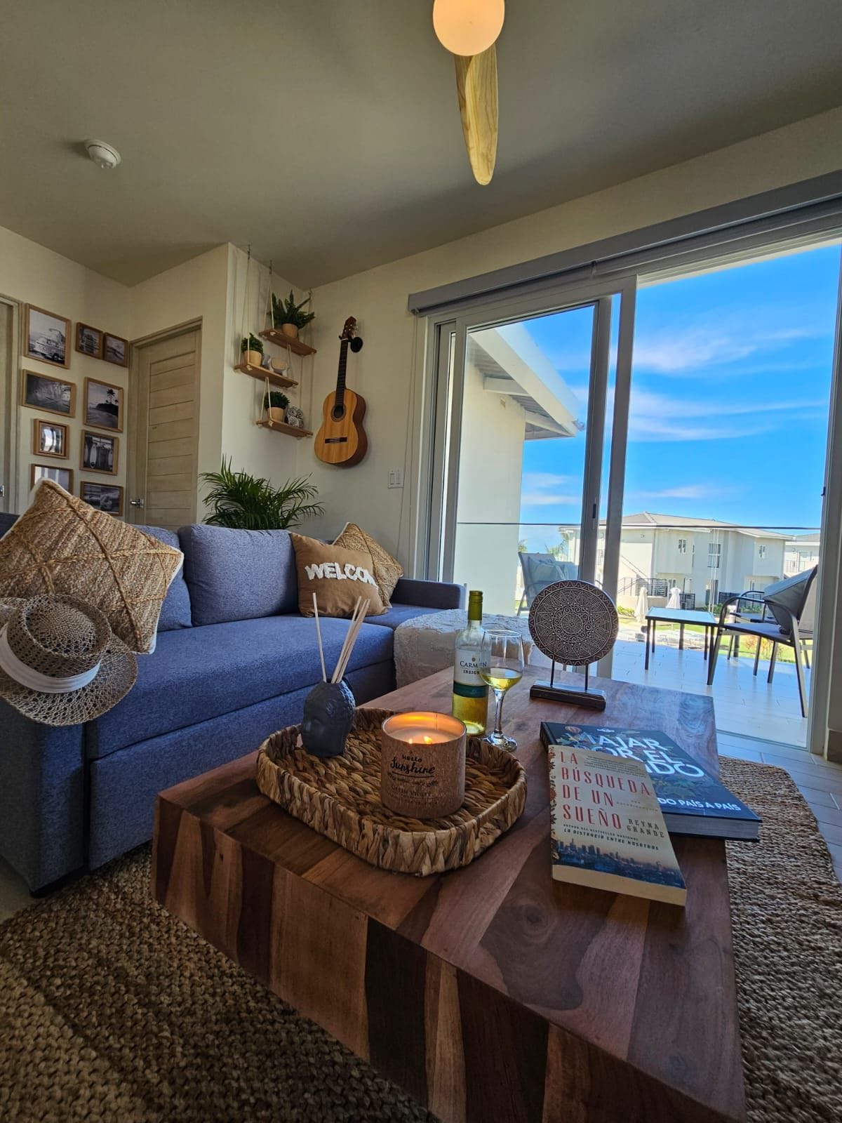 A cozy living area features a comfortable blue couch adorned with decorative pillows. A wooden coffee table, holding a candle and a book, is centrally placed. Natural light fills the space, highlighted by sliding glass doors that open to a patio with outdoor seating.