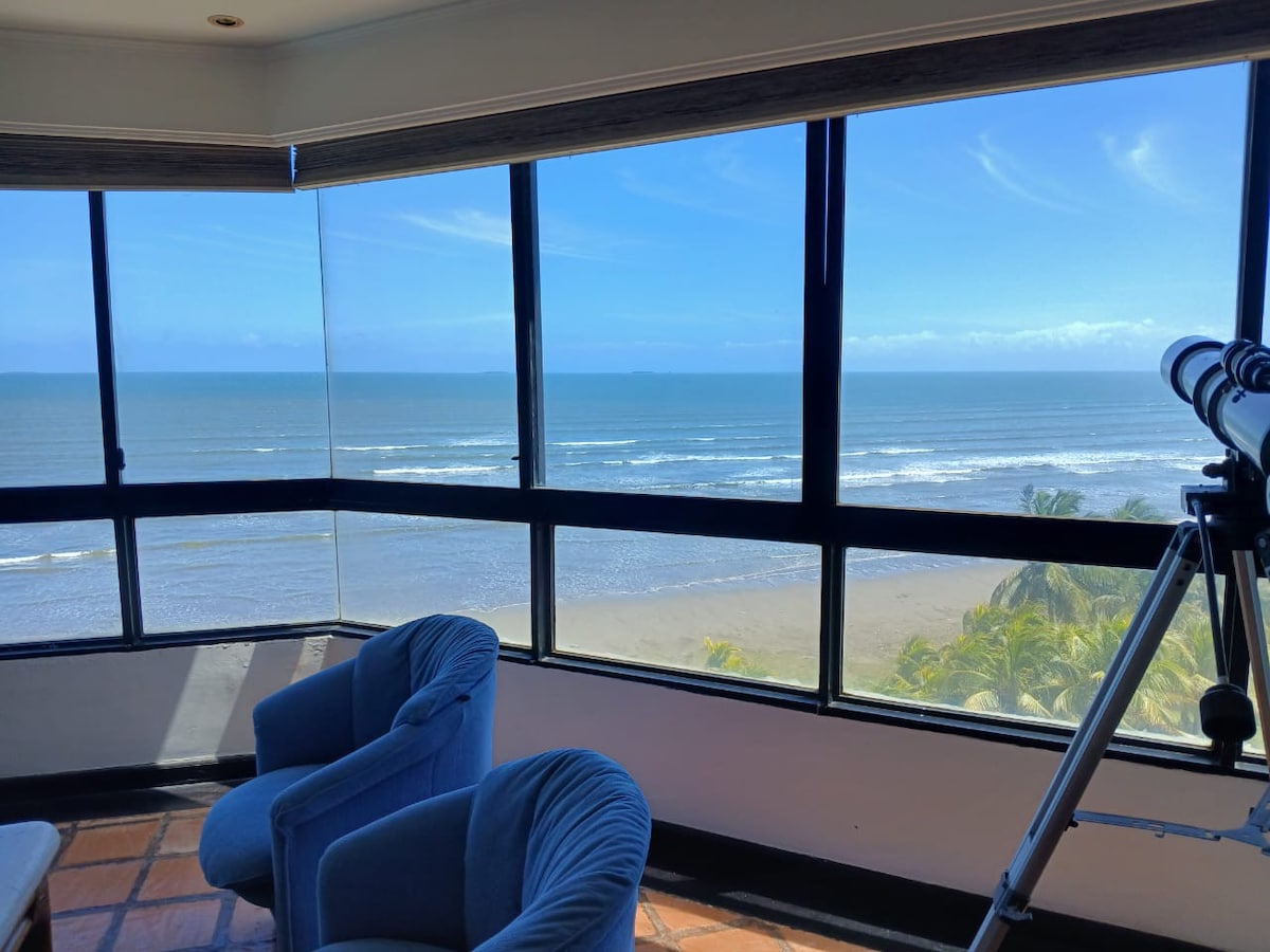 A bright and inviting living space features large windows offering a panoramic view of the ocean and beach. Two plush blue chairs are positioned to enjoy the scenery, while a telescope is available for closer observation of the coastal landscape.