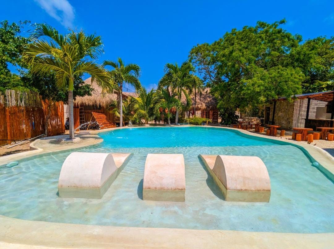 A large outdoor swimming pool is framed by lush palm trees and green foliage. White, curved seating areas are integrated into the water, allowing for relaxation. Surrounding lounge chairs and wooden tables create a serene environment for family gatherings.