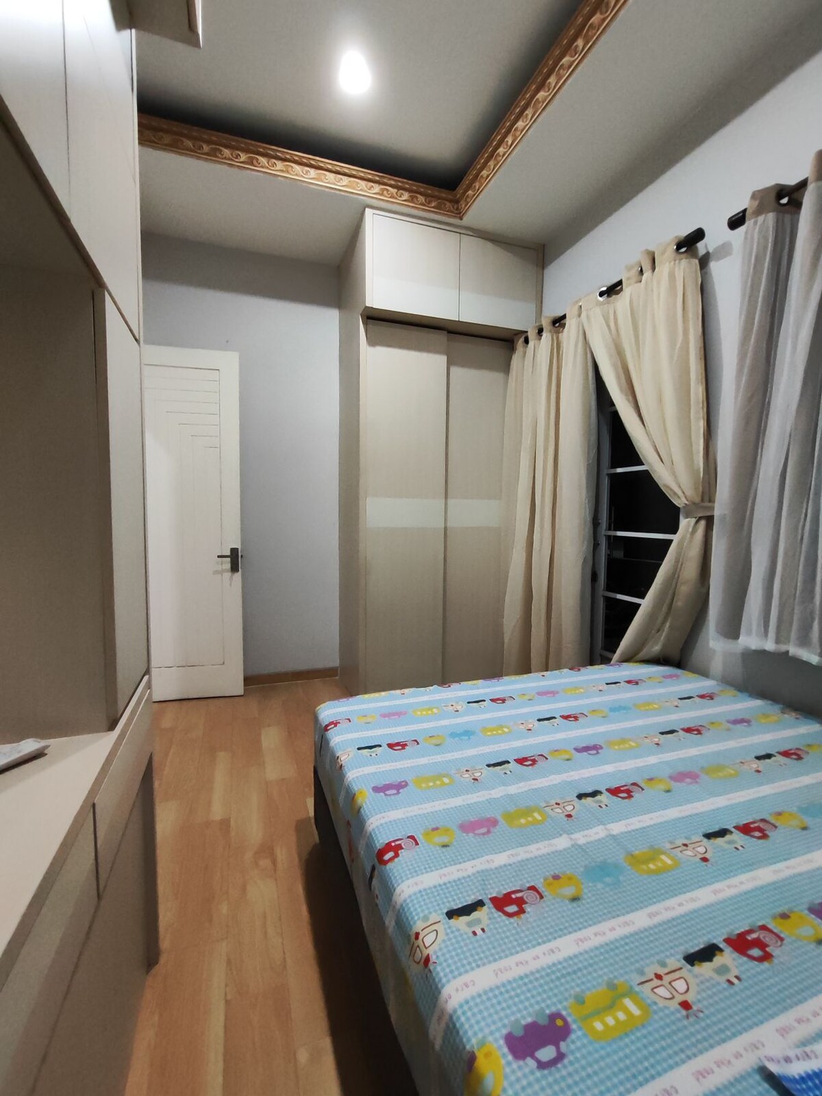 A bedroom featuring a spacious bed with a colorful patterned comforter. A wooden closet with sliding doors is placed against one wall. Light-colored curtains frame the window, and the room is complemented by wooden flooring and subtle ceiling details.