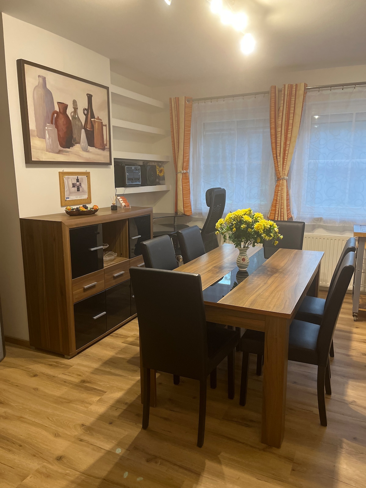 A modern dining area features a wooden table surrounded by six black chairs. A sideboard presents a mix of glass and wood finishes. A painting of vases adorns the wall, while natural light filters in through curtains, enhancing the inviting space.