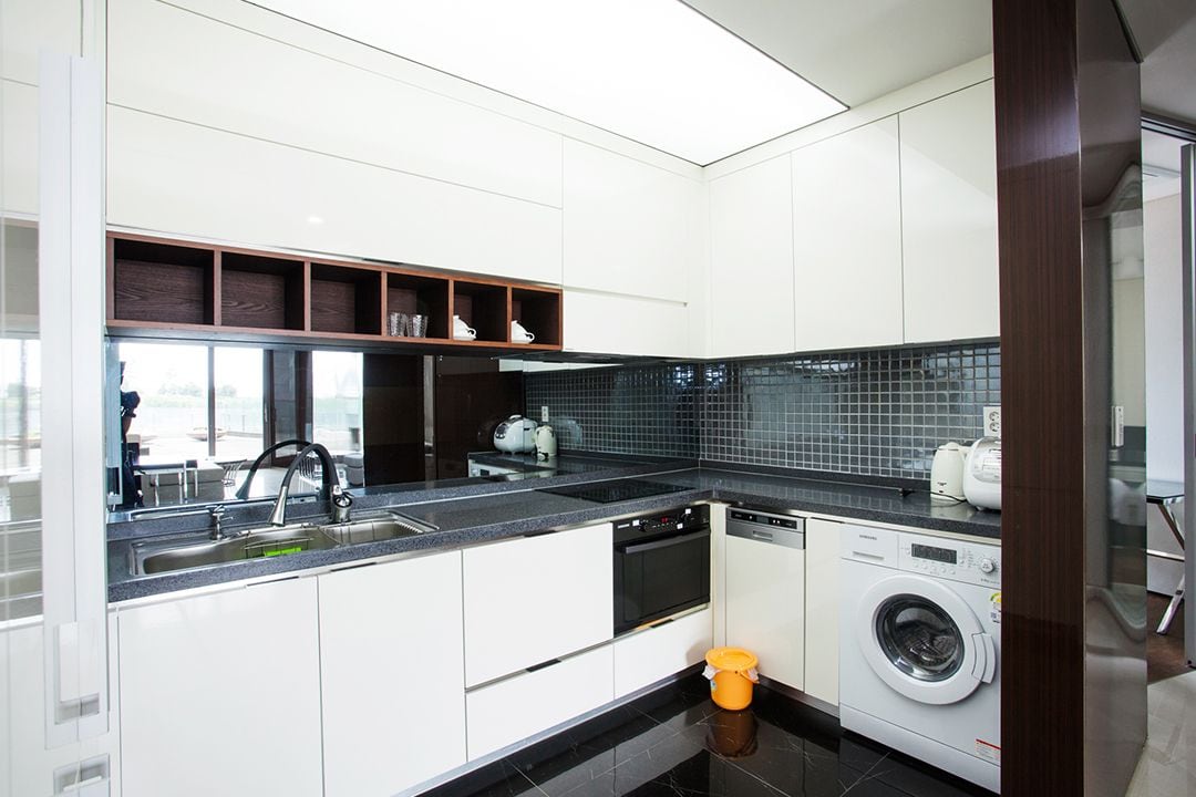 A modern kitchen features sleek, white cabinetry and a dark countertop. Stainless steel appliances, including a dishwasher and a washing machine, are present. A mosaic tile backsplash adds texture, while ample storage space is provided by open shelving above. Bright overhead lighting is available.