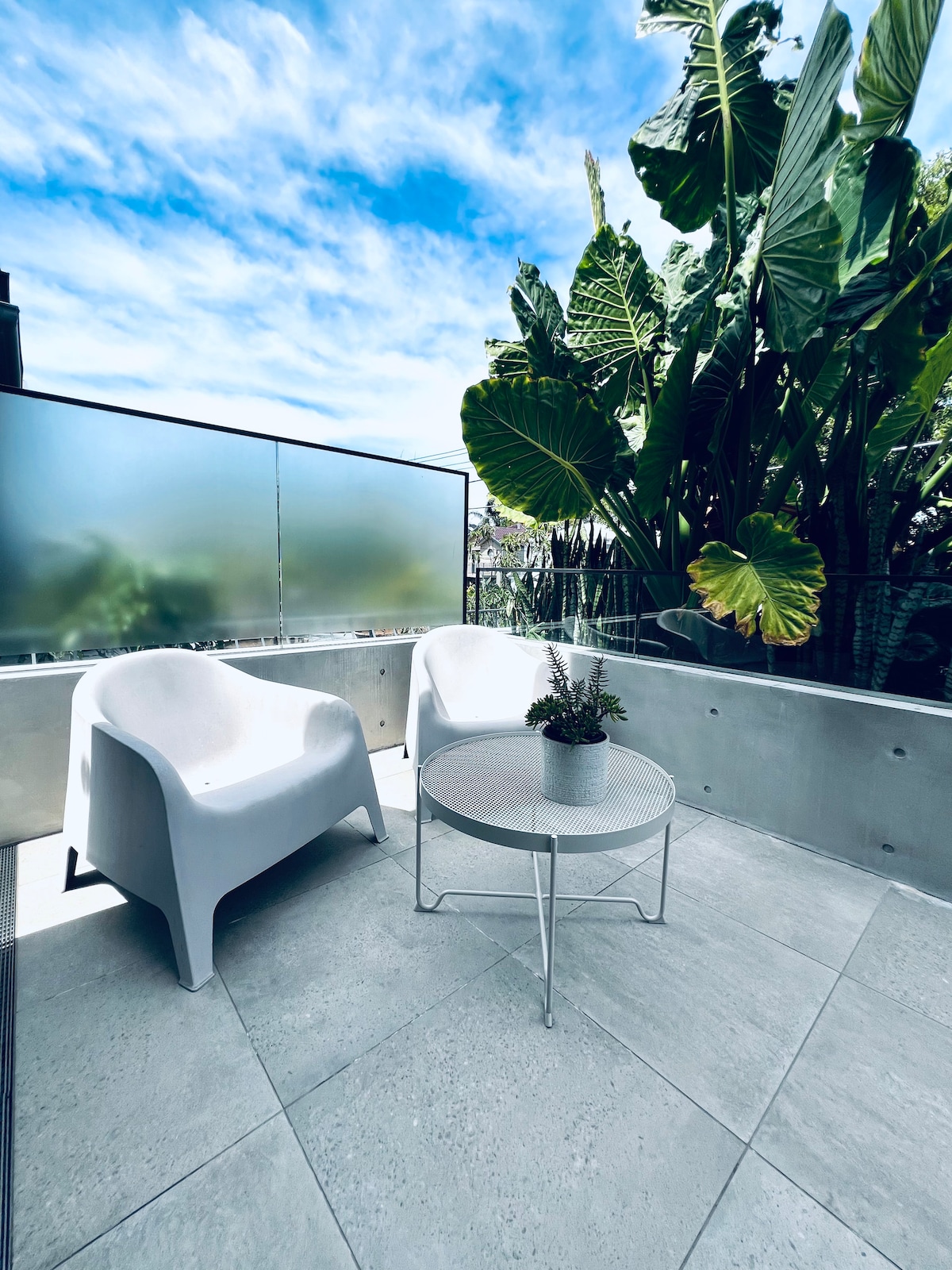 An outdoor terrace is presented with two sleek white chairs and a small round table. Tropical plants surround the area, providing a lush backdrop. The flooring is composed of light gray tiles, and a low wall offers partial privacy while still allowing natural light.