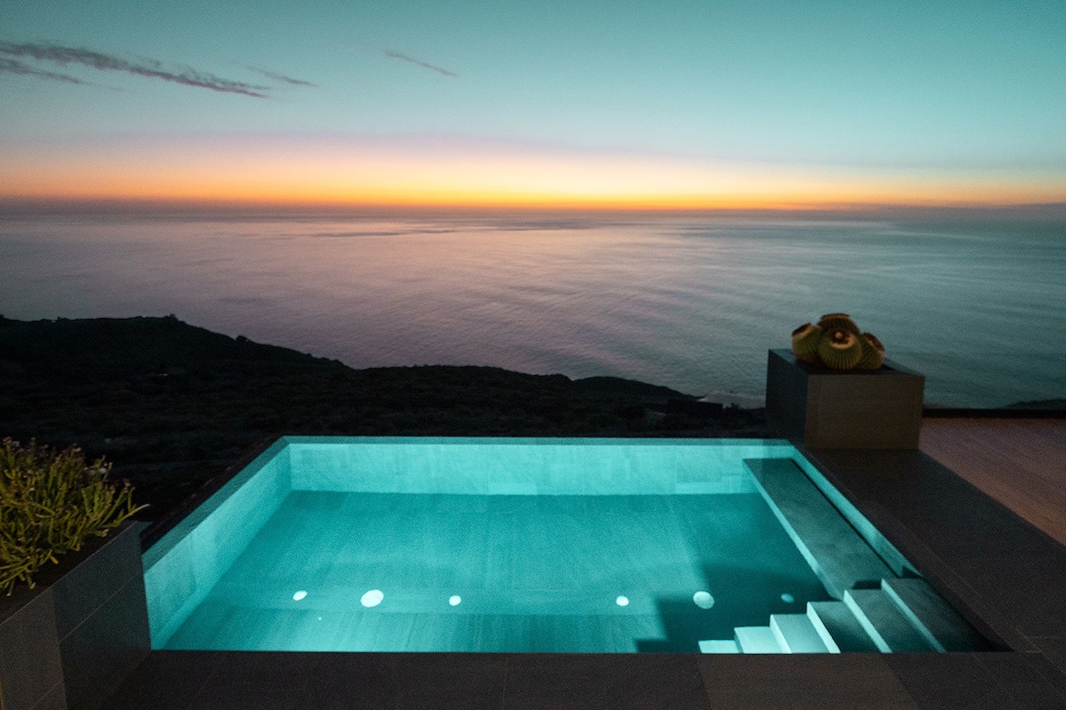 An infinity pool is depicted, showcasing a serene turquoise hue against the backdrop of a vibrant sunset over the Atlantic Ocean. The surroundings are characterized by smooth stone steps leading into the pool and a gentle glow reflecting off the water.