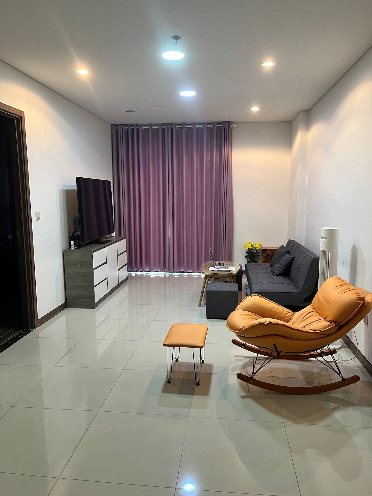 A living room area features a soft gray sofa and an inviting yellow rocking chair. A small side table accompanies the chair, while a television is mounted on a light gray stand. Natural light filters through sheer purple curtains, accompanied by decorative plants.