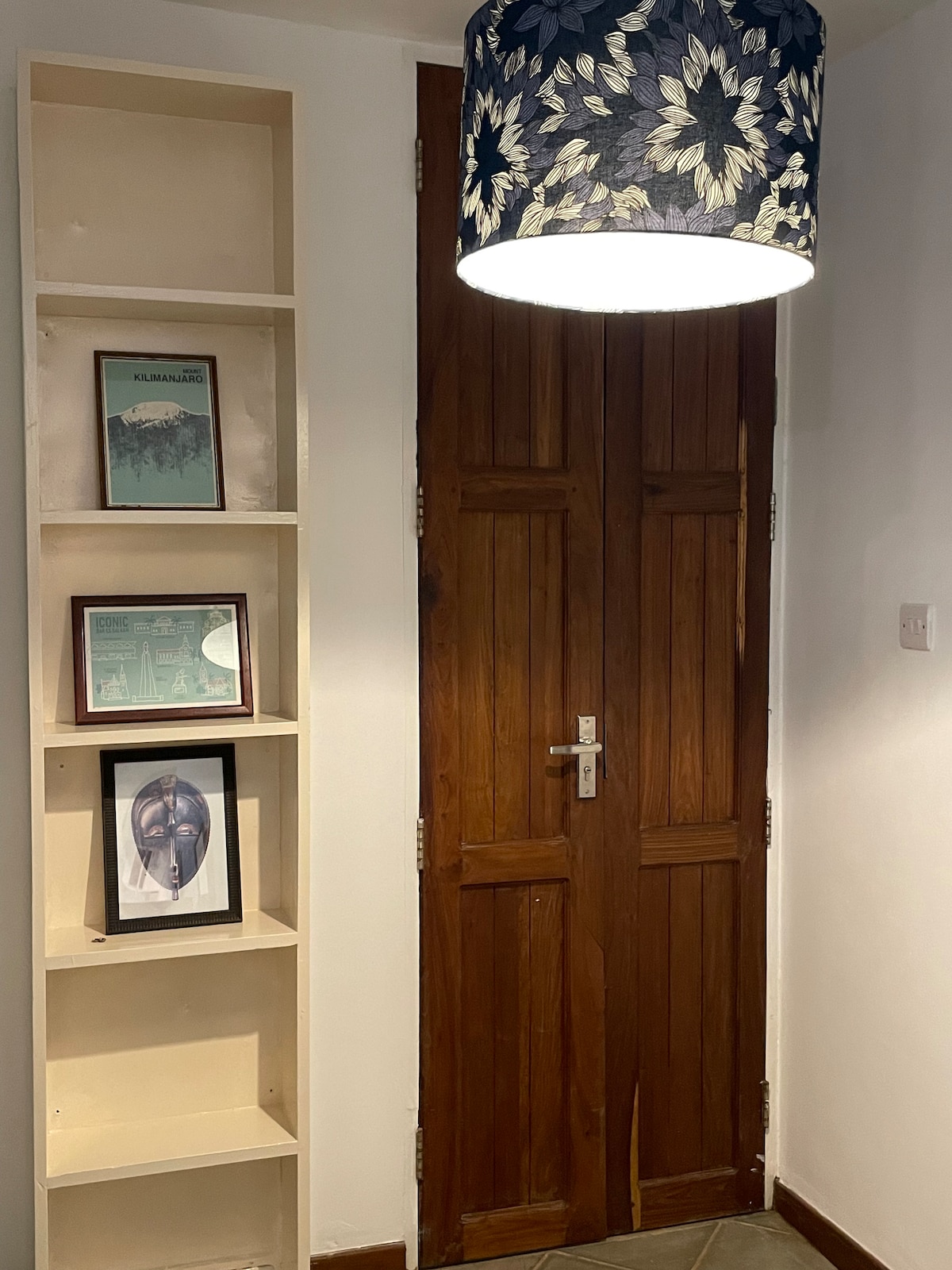 A wooden door is featured prominently in the image, surrounded by a well-lit entryway. A shelving unit holds framed photographs and artwork, adding character. A decorative lamp with a floral design provides soft illumination, enhancing the inviting atmosphere of the entrance.