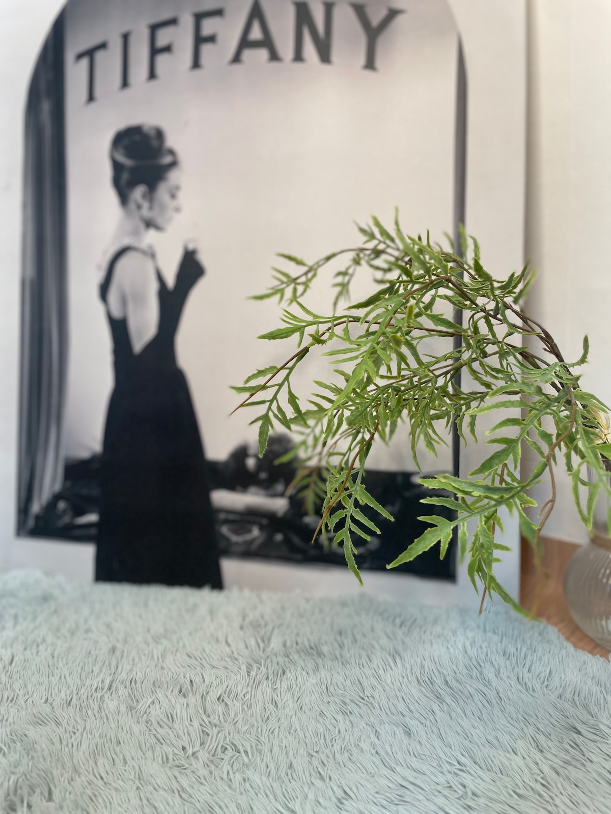 A decorative element features a small green plant beside a soft blue textured surface. In the background, a black and white poster displays the word 'TIFFANY' along with an elegant figure in a black dress, contributing to a stylish and sophisticated atmosphere.