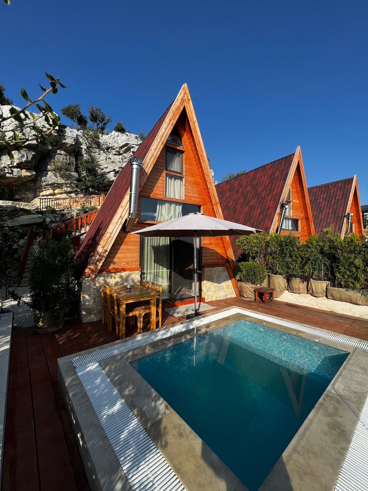 Three A-shaped bungalows are situated against a backdrop of rocky terrain. A private swimming pool is visible in the foreground, accompanied by outdoor seating set under a patio umbrella. Lush greenery and a wooden deck enhance the tranquil outdoor space.