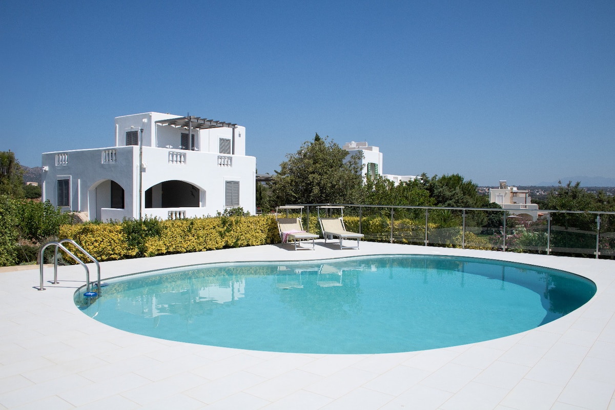 A private circular swimming pool reflects the clear blue sky, surrounded by a spacious terrace. Lush green gardens provide a backdrop, while white-walled villas are visible in the distance. Sun loungers are positioned nearby, inviting relaxation in the sun.