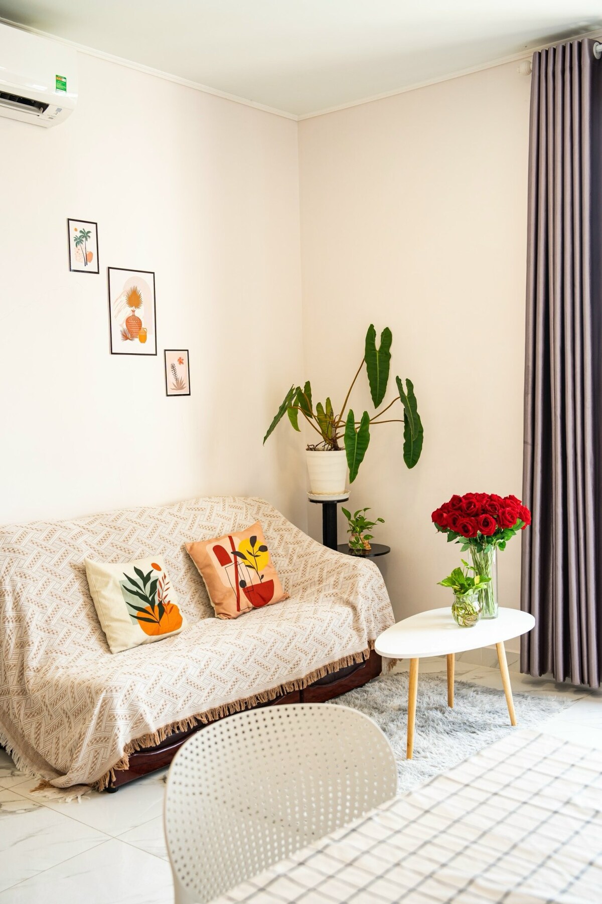 A cozy sitting area is highlighted by a sofa adorned with patterned throw blankets and decorative cushions. A small round table holds a vibrant bouquet of red roses, while wall art featuring various designs adds a personal touch to the space. Green plants are positioned nearby, enhancing the setting.