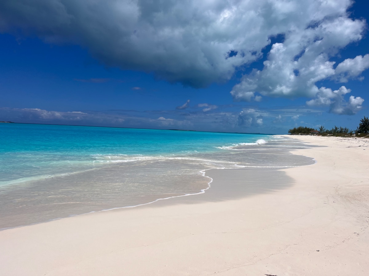 A serene beach scene showcases soft, white sand meeting clear turquoise waters under a partly cloudy sky. Gentle waves lap at the shore, and a small stretch of lush greenery is visible in the distance, offering a peaceful coastal view.