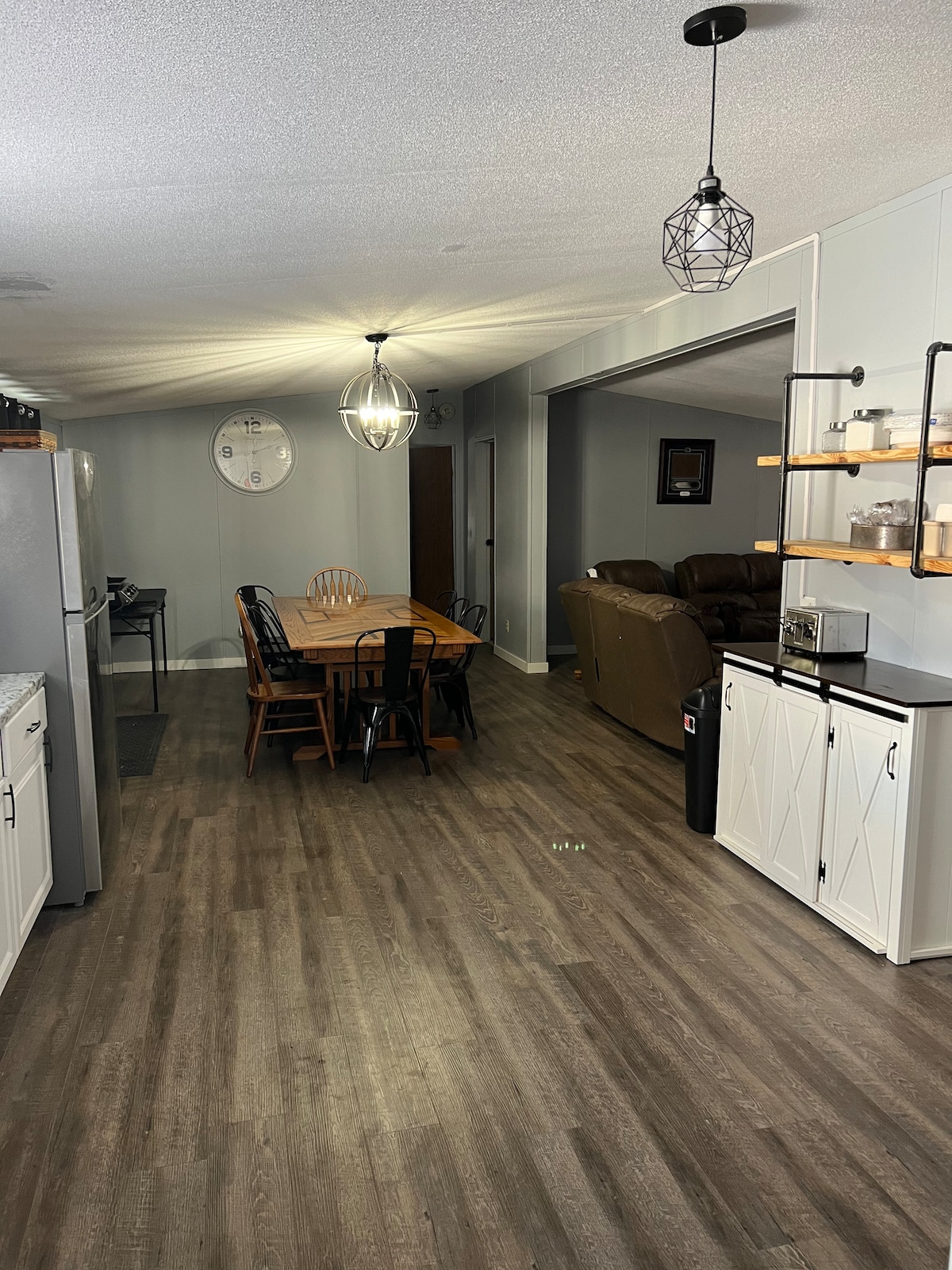 An open floor plan showcases a dining area with a wooden table surrounded by chairs. Soft lighting is provided by pendant lamps above. The cozy sitting area features a brown couch in the background, while light-colored cabinets offer storage in the foreground.