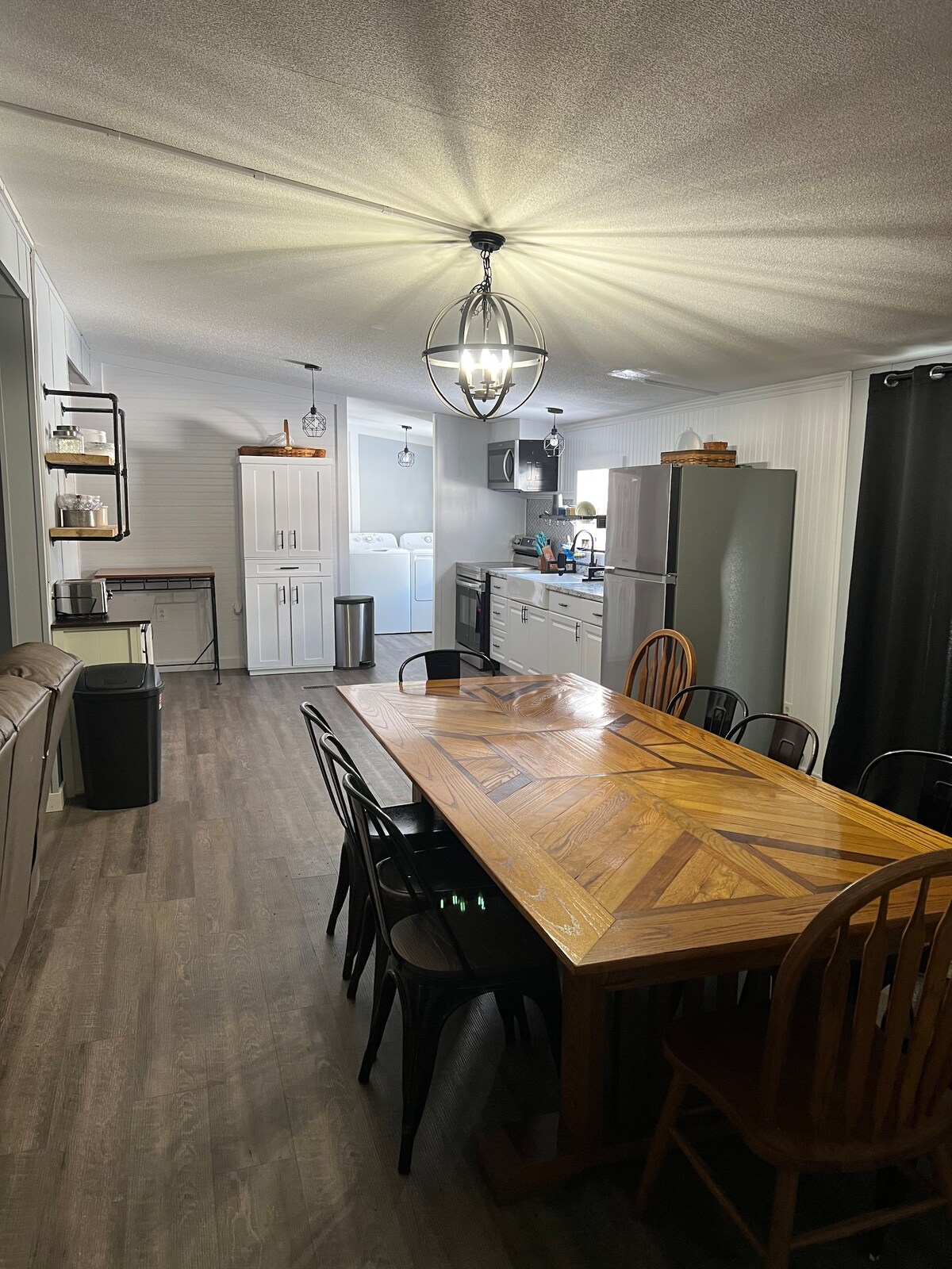 A spacious dining area features a large wooden table surrounded by a mix of black and wooden chairs. A modern light fixture hangs above, illuminating the area. The kitchen is visible in the background, equipped with stainless steel appliances and cabinetry.