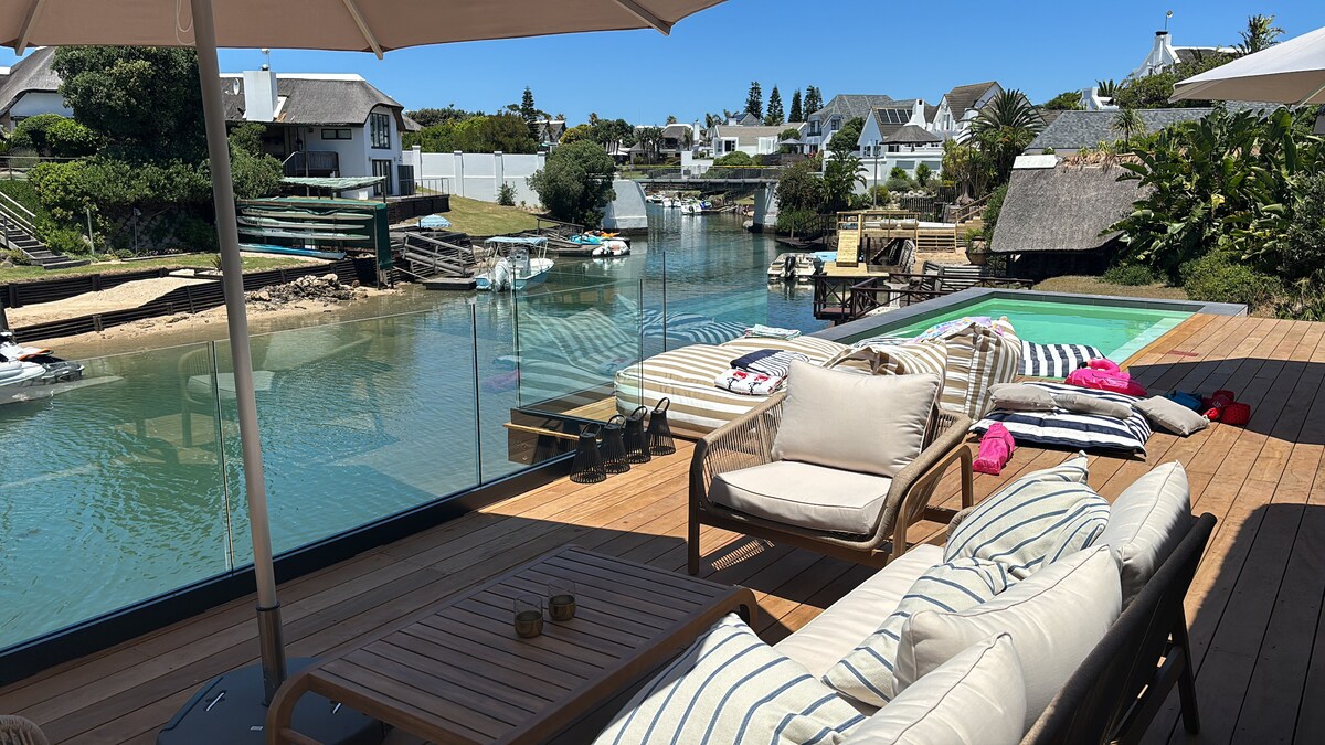 A spacious deck provides views of the tranquil canal, featuring a heated swimming pool and comfortable seating. Light-colored lounge chairs are arranged around a wooden coffee table. Umbrellas offer shade, while nearby boats are moored, enhancing the peaceful outdoor setting.