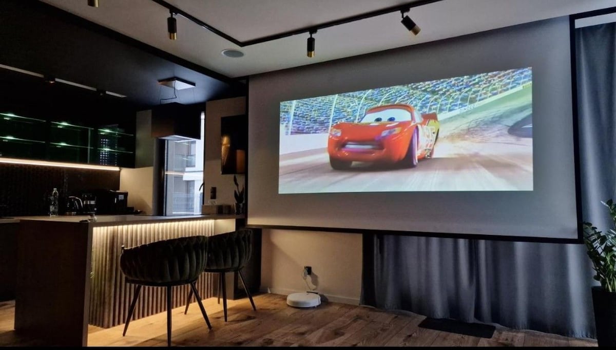 A projector screen displays a vibrant scene from an animated film, creating a home cinema experience. A modern kitchen area is visible in the background, featuring sleek cabinetry and four stylish chairs at the island. Soft lighting adds to the contemporary design.