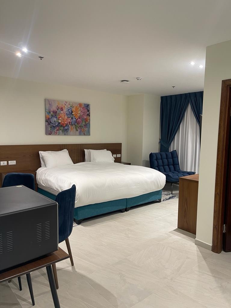A spacious studio features a king-size bed with white bedding and accent pillows. Two blue chairs are positioned near a compact kitchenette. A colorful floral artwork adorns the wall above the bed, while large windows are framed by blue curtains, allowing soft light to enter.