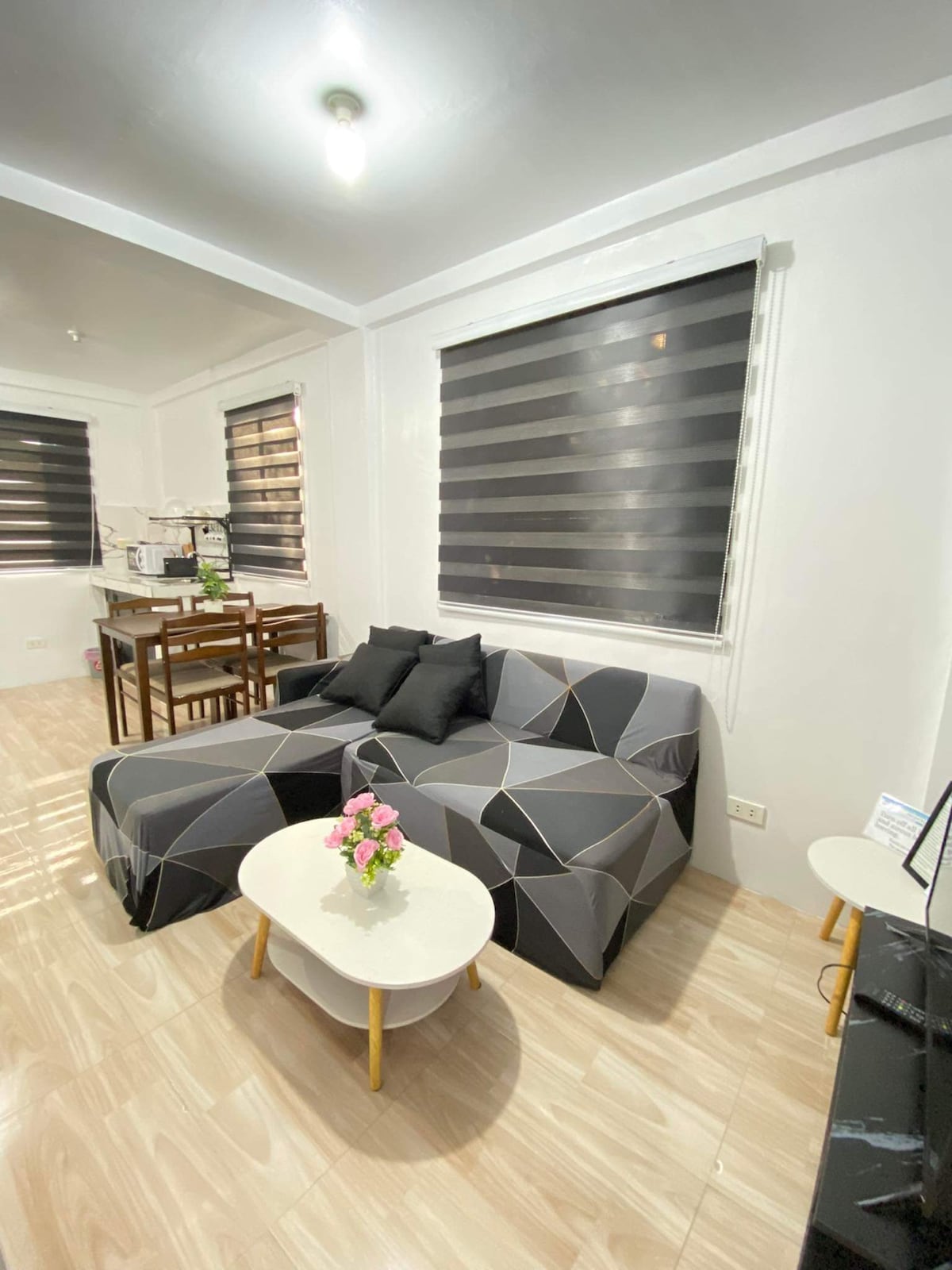 A minimalist living area features a black and gray sectional sofa with accent pillows, positioned next to a small white coffee table adorned with flowers. Large windows with dark blinds allow natural light to brighten the space, complementing the light-colored flooring.