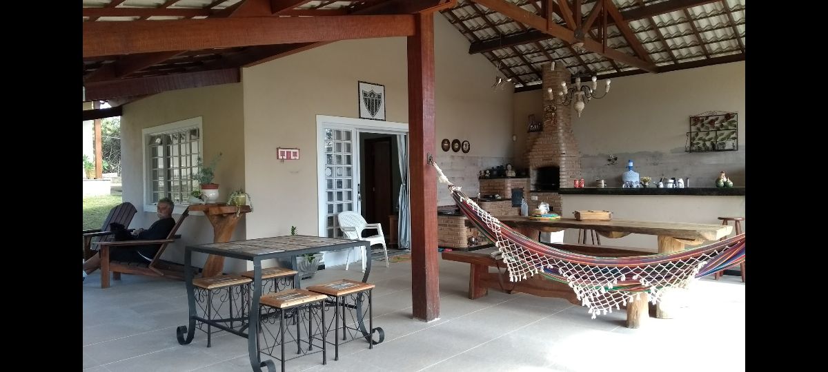 An open-concept living space features a dining table surrounded by chairs and a hammock, offering a relaxed atmosphere. A kitchen area is visible with various appliances. Natural light illuminates the room through large windows, enhancing the connection to the outdoors.