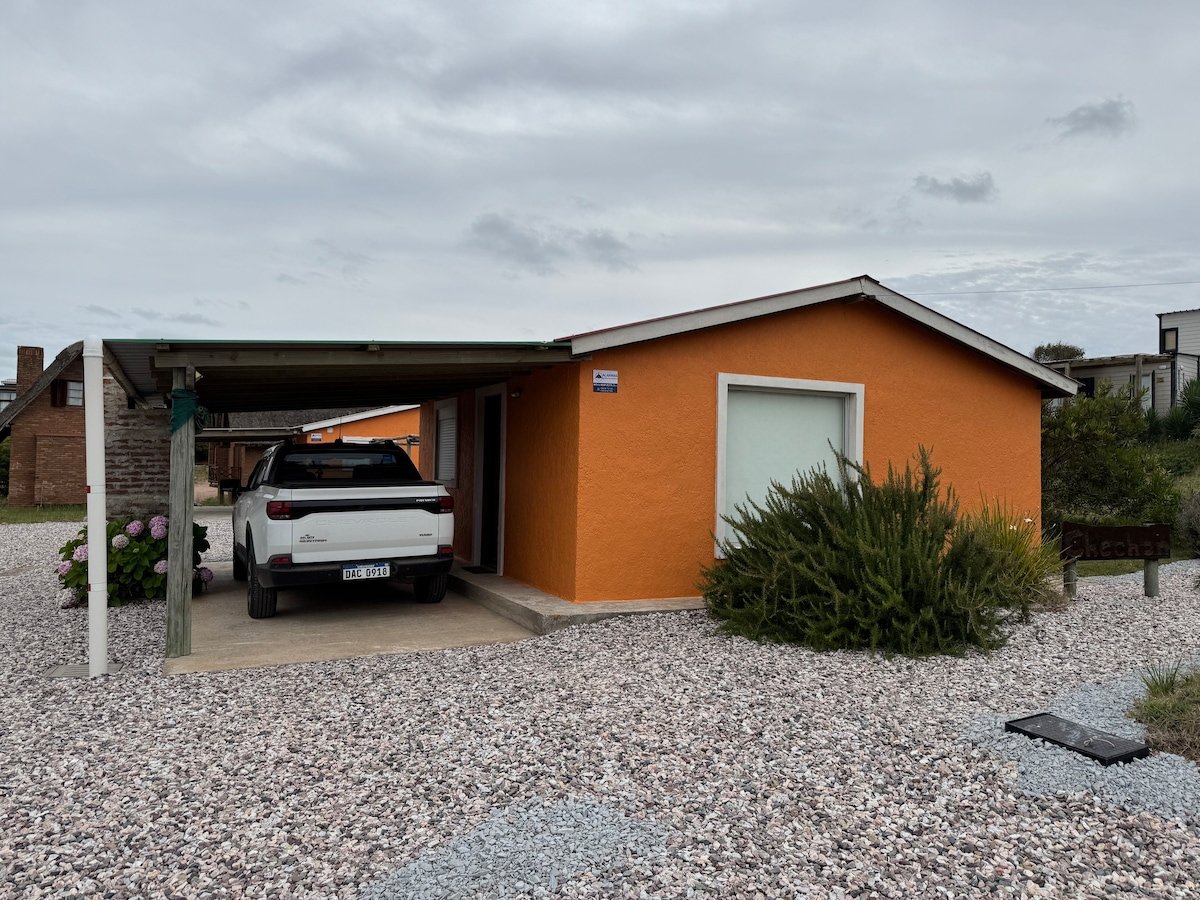 A single-story orange house is set amidst a gravel driveway, featuring a carport partially shaded by a roof. Green shrubs and flowering plants are visible, adding greenery to the exterior. The entrance has a large window allowing natural light into the space.