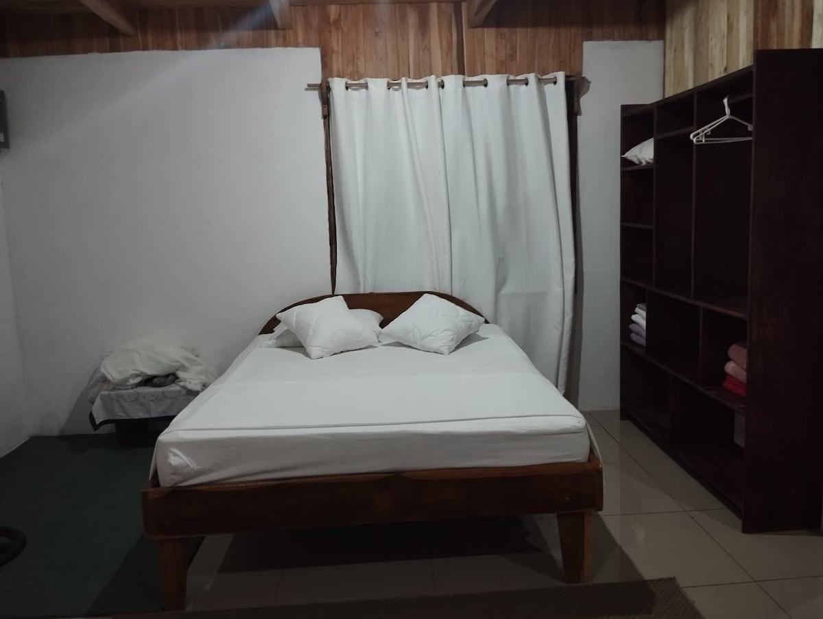 A simple sleeping area features a wooden bed with two white pillows atop a spacious mattress. A neatly organized shelf holds folded towels, while a light-colored curtain partially covers the window, allowing natural light to illuminate the room.