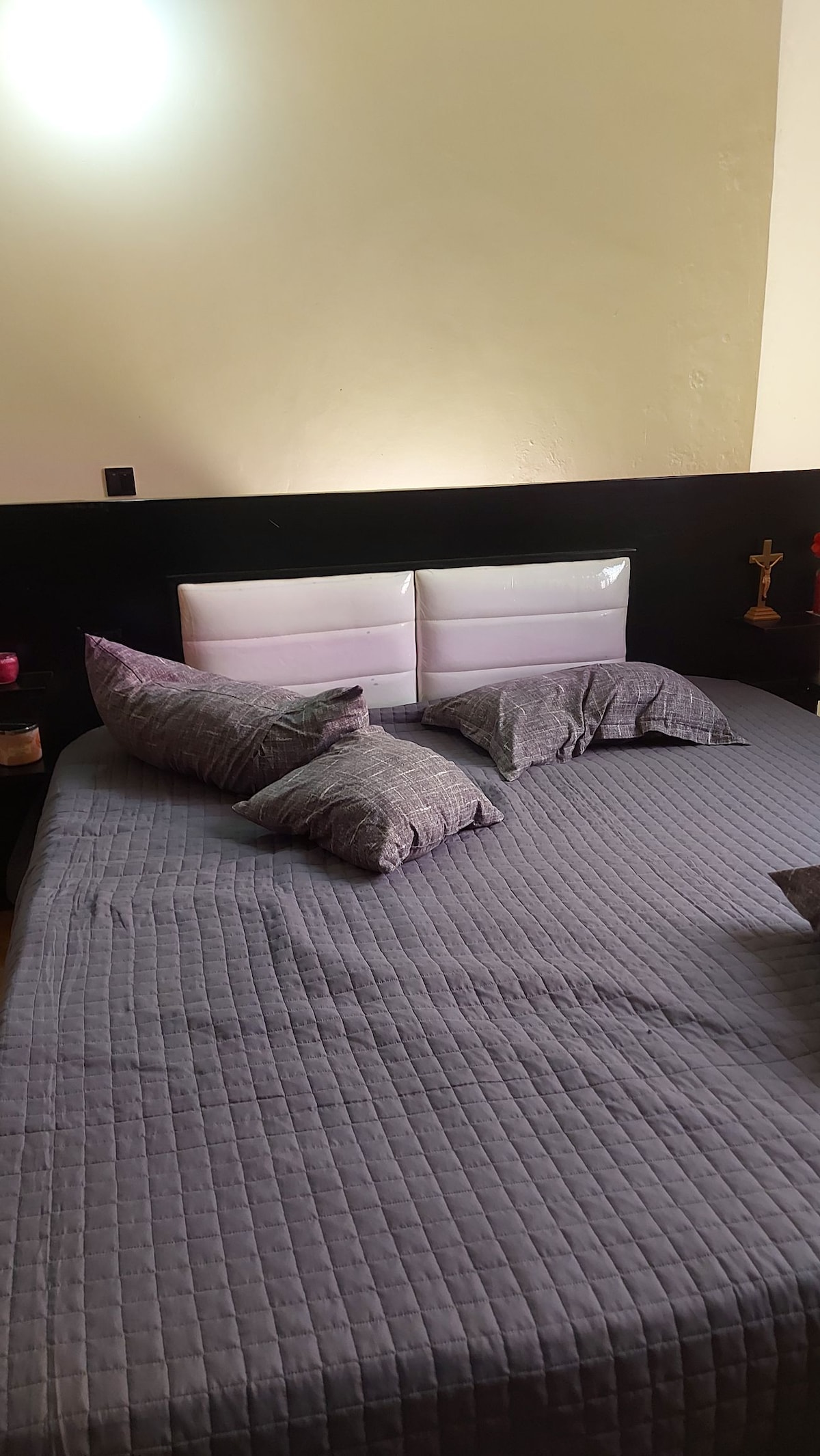 A spacious bed is featured with a grey quilted cover and several scattered pillows. The headboard is designed with sleek white panels complementing the dark wall behind. Soft lighting is provided from a wall-mounted fixture above the bed.