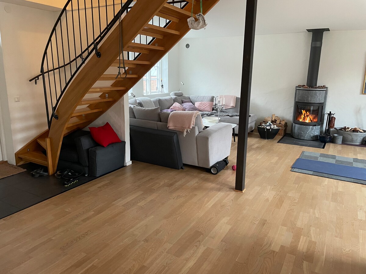 A spacious living area features a wooden staircase leading to an upper level. A cozy seating arrangement with light-colored sofas is positioned near a stove. Soft throws and cushions add comfort to the space, while a muted color palette creates a calming ambiance.