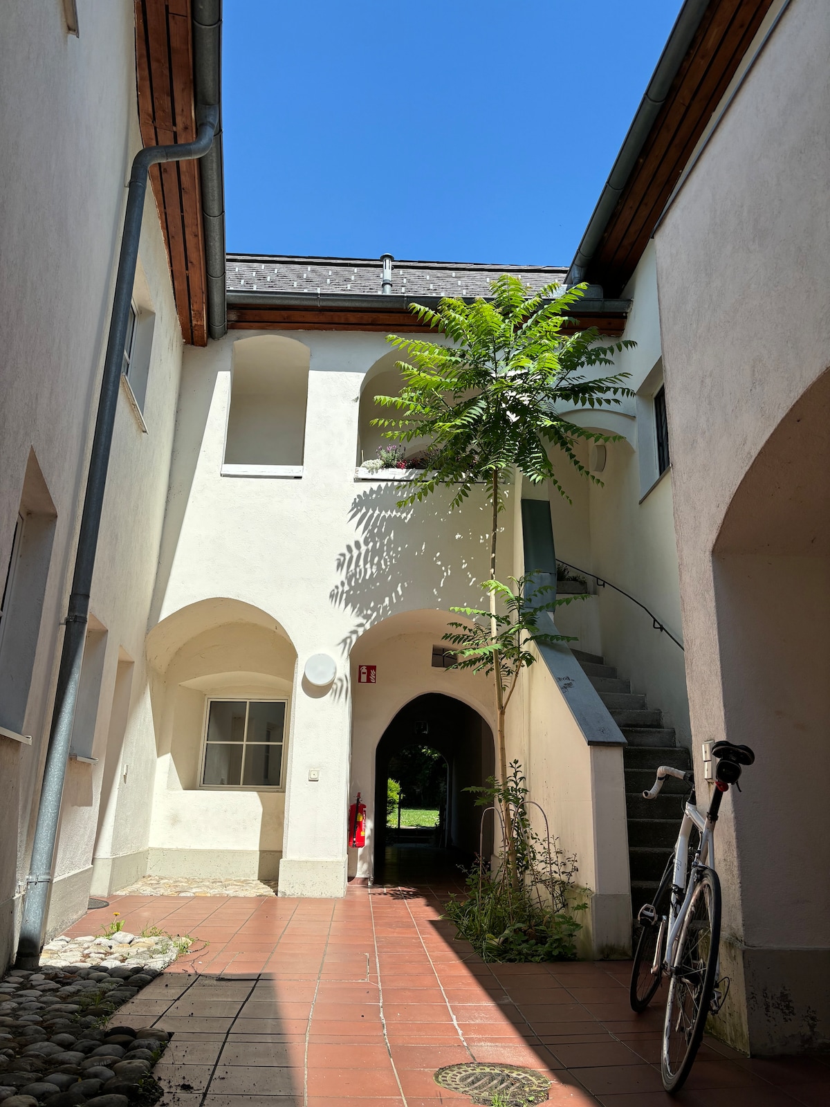 An open courtyard is featured, framed by white walls and blue skies overhead. A narrow staircase leads to an upper level, while a bicycle is parked beside it. A tall tree adds greenery to the space, creating a serene atmosphere.