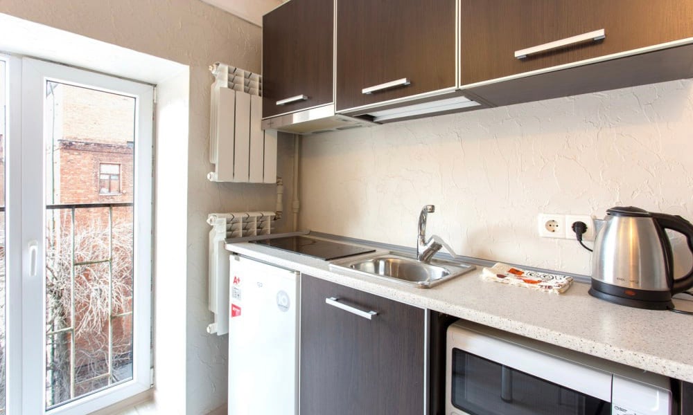 The kitchen area features dark wood cabinets and a fitted countertop. A stainless steel kettle and a small microwave are visible, alongside a sink. Natural light enters through a window, offering a view of the outside, while heating radiators are mounted on the wall.