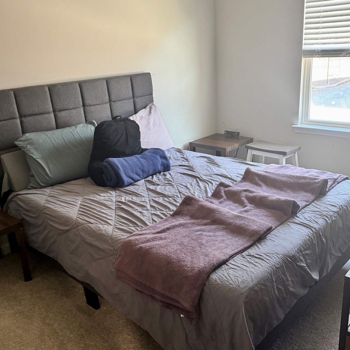 A spacious bed with a grey upholstered headboard is positioned against a light-colored wall. Soft linens in muted tones are spread across the bed. A couple of pillows are arranged at the head, and a small nightstand with a simple lamp is placed beside it. Natural light filters in from the window.