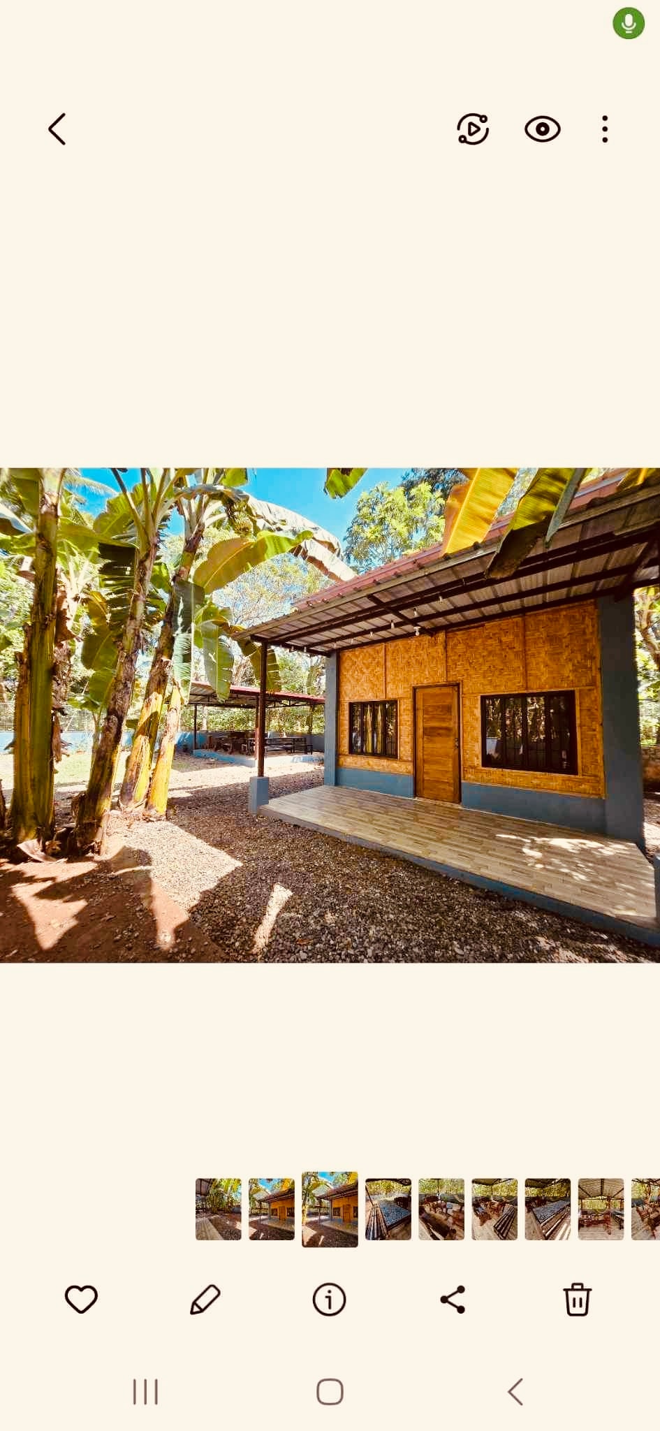 A charming exterior of a single-story house is framed by lush banana trees. The structure features wooden accents and large windows allowing ample natural light. A wooden deck extends along the front, providing an inviting entrance shaded by the overhanging roof.